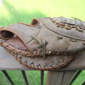 Peut inclure: Un gant de baseball vintage, usé, dans des tons de beige et de marron. Le gant montre des signes d'utilisation avec des coutures et des lacets en cuir visibles. Le gant repose sur une surface en bois, suggérant une utilisation en extérieur.