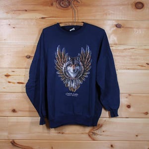 May include: A navy blue sweatshirt with a wolf design. The wolf has a detailed face with grey and white fur, and is surrounded by brown and gold feathered wings. The text "Curve Lake Indian Reserve" is below the wolf image. The sweatshirt hangs on a wooden background.