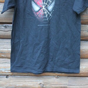 May include: A black short-sleeve t-shirt featuring a graphic of the Spider-Man mask with a red and black web pattern on the upper chest. The text Spider-Man is printed in small white letters directly below the mask graphic on the front of the shirt.