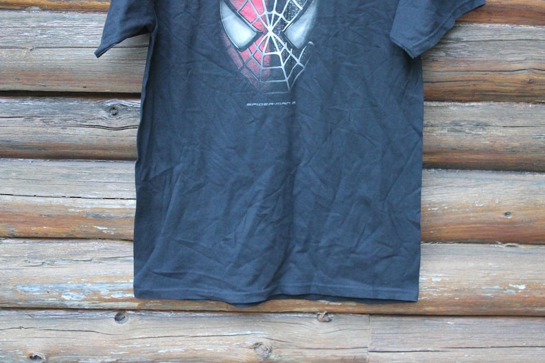 May include: A black short-sleeve t-shirt featuring a graphic of the Spider-Man mask with a red and black web pattern on the upper chest. The text Spider-Man is printed in small white letters directly below the mask graphic on the front of the shirt.