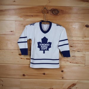 May include: White hockey jersey with blue and white stripes on the sleeves. The jersey has a blue maple leaf logo with "Toronto Maple Leafs" text on it.