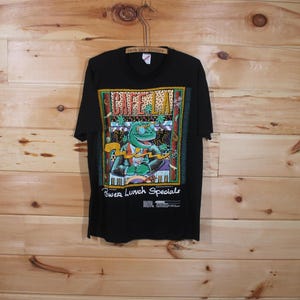 May include: Black t-shirt featuring a colourful graphic of a cartoon lizard playing a keyboard. The graphic includes the text "Cafe La" and "Power Lunch Specials".