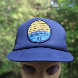 Puede incluir: Gorra de camionero azul marino con un parche que representa una puesta de sol sobre el agua, con el texto "Darlington G.S. 87". La gorra tiene una visera curva y una parte trasera de malla negra.