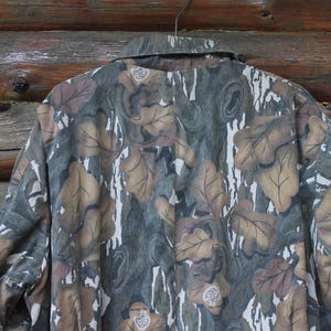 Puede incluir: Chaqueta de camuflaje con un estampado de hojas en tonos marr&oacute;n, verde y gris. La chaqueta est&aacute; colgada de una percha met&aacute;lica sobre un fondo de madera. El cuello es visible.