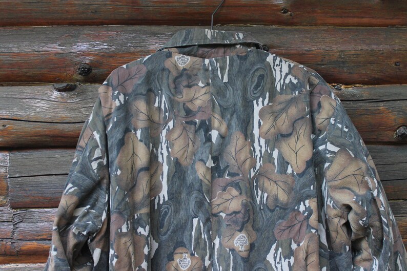 Puede incluir: Chaqueta de camuflaje con un estampado de hojas en tonos marr&oacute;n, verde y gris. La chaqueta est&aacute; colgada de una percha met&aacute;lica sobre un fondo de madera. El cuello es visible.