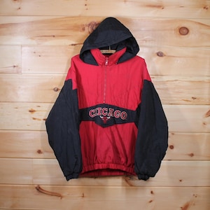 Puede incluir: Una chaqueta roja y negra con capucha, cierre de cremallera y cordón de ajuste en la cintura. La chaqueta tiene un logotipo de los Chicago Bulls y la palabra "CHICAGO" impresa en la parte delantera.