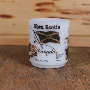 Może przedstawiać: Biały kubek ceramiczny z czarno-złotym wzorem przedstawiającym flagę Nowej Szkocji oraz napis "Nova Scotia" i "Canada". Kubek zawiera również napis "Peggy's Cove" i "Entrance Citadel Hill Halifax".