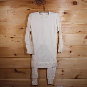 May include: Cream thermal underwear set, comprising a long-sleeved top and matching trousers. The top has a crew neck and ribbed cuffs, and the trousers have ribbed cuffs at the ankles. The set is hanging on a vintage metal hanger against a wooden background.