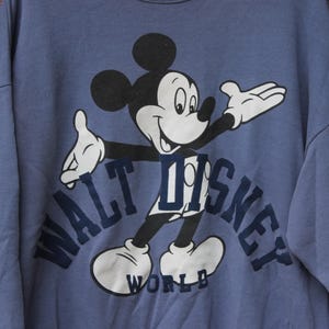 Puede incluir: Sudadera azul con un dise&ntilde;o de Mickey Mouse. El texto "WALT DISNEY WORLD" en azul marino. La sudadera tiene cuello redondo. La imagen est&aacute; sobre un fondo de madera.
