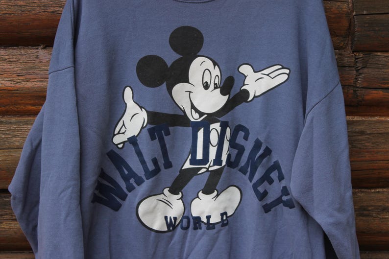 Puede incluir: Sudadera azul con un dise&ntilde;o de Mickey Mouse. El texto "WALT DISNEY WORLD" en azul marino. La sudadera tiene cuello redondo. La imagen est&aacute; sobre un fondo de madera.