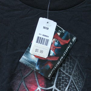 May include: A black t-shirt featuring a Spider-Man 3 graphic print. A white retail tag is attached to the collar, displaying the size M/M, a barcode, SKU 609804, and a price of $5.99. The tag also shows a partial image of the Spider-Man suit.
