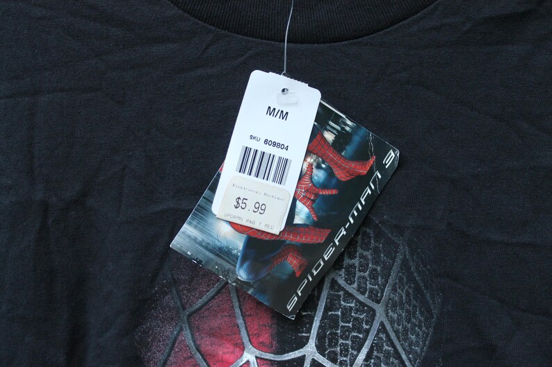 May include: A black t-shirt featuring a Spider-Man 3 graphic print. A white retail tag is attached to the collar, displaying the size M/M, a barcode, SKU 609804, and a price of $5.99. The tag also shows a partial image of the Spider-Man suit.