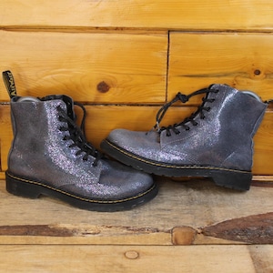 Puede incluir: Un par de botas de combate grises con purpurina, cordones negros y costuras amarillas. Las botas están hechas de un material brillante y texturizado y tienen un diseño clásico con cordones. Las botas se exhiben sobre una superficie de madera.