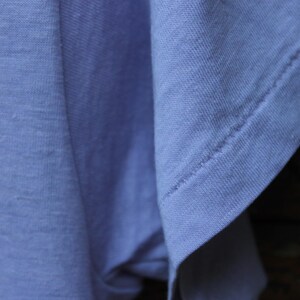 May include: A light blue t-shirt featuring a short sleeve with visible double-stitched hem detailing. The fabric has a soft, matte texture and a consistent solid color throughout the garment, showcasing the sleeve construction and shoulder seam.