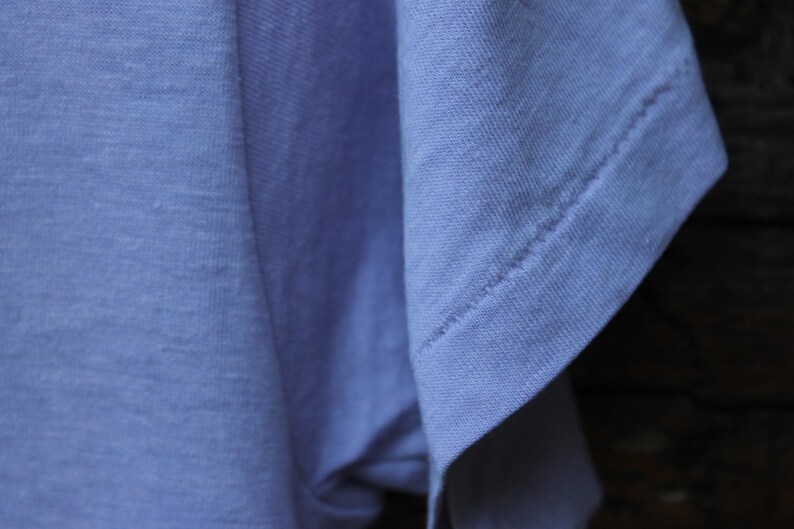 May include: A light blue t-shirt featuring a short sleeve with visible double-stitched hem detailing. The fabric has a soft, matte texture and a consistent solid color throughout the garment, showcasing the sleeve construction and shoulder seam.