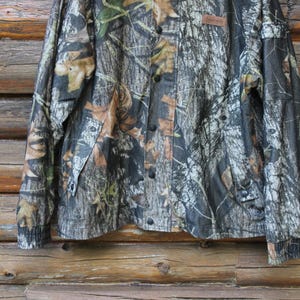 May include: Camouflage button-up shirt with long sleeves. The shirt features a pattern of brown, green, and tan leaves and branches. The shirt has black buttons and a small brown tag. The shirt is hanging against a wooden background.