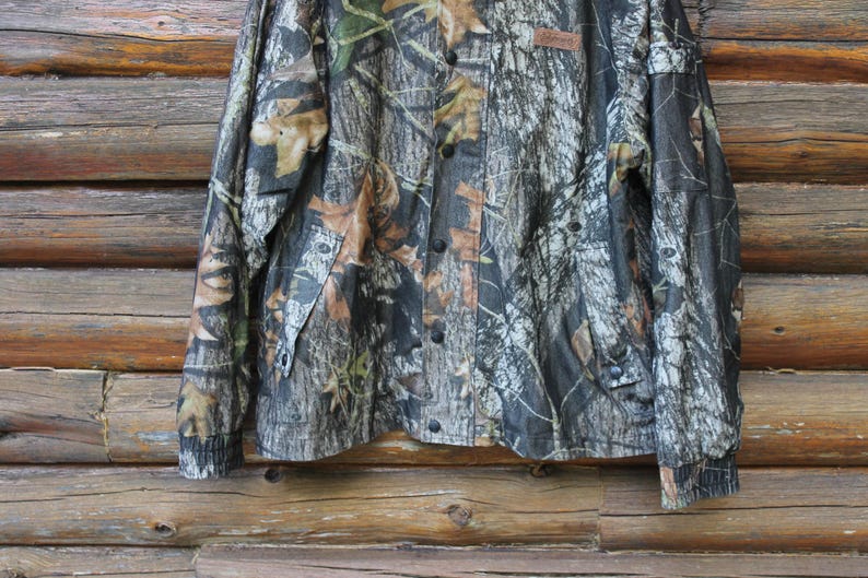 May include: Camouflage button-up shirt with long sleeves. The shirt features a pattern of brown, green, and tan leaves and branches. The shirt has black buttons and a small brown tag. The shirt is hanging against a wooden background.
