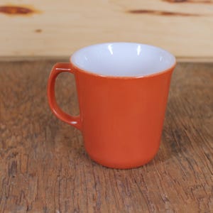 Puede incluir: Una taza de cerámica de color terracota con interior blanco. La taza tiene un asa redondeada y un borde ligeramente acampanado.