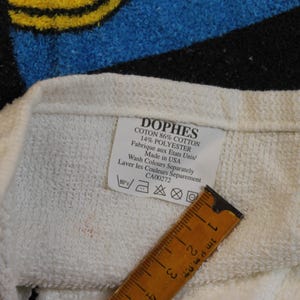 May include: Close-up of a white towel with a label that reads "DOPHES COTON 86% COTTON 14% POLYESTER Made in USA." A wooden ruler is placed on the towel. The towel has blue, black, and yellow accents.