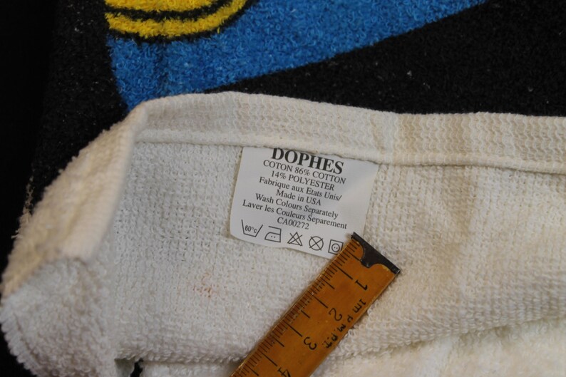 May include: Close-up of a white towel with a label that reads "DOPHES COTON 86% COTTON 14% POLYESTER Made in USA." A wooden ruler is placed on the towel. The towel has blue, black, and yellow accents.
