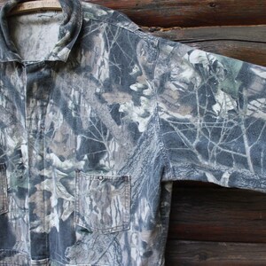 May include: Camouflage jacket featuring a pattern of green, brown, and grey, with two front pockets and a collared neckline. The jacket's fabric mimics a forest environment. It is hanging on a wooden hanger.