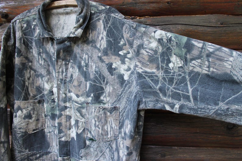 May include: Camouflage jacket featuring a pattern of green, brown, and grey, with two front pockets and a collared neckline. The jacket's fabric mimics a forest environment. It is hanging on a wooden hanger.
