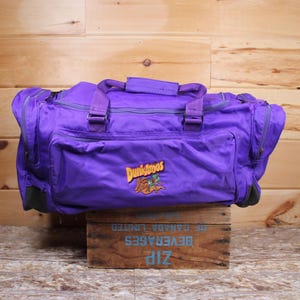 May include: Purple duffel bag with a Dunkaroos logo featuring a cartoon bear and the text "Dunkaroos".