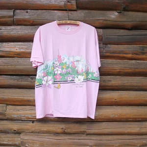 May include: Pink t-shirt with a floral print and a butterfly. The print features a variety of flowers and a hummingbird. The text on the shirt reads "Prince George BC Canada".