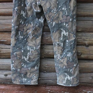 Puede incluir: Pantalones de camuflaje con un estampado de hojas en tonos marr&oacute;n, gris y blanco. Los pantalones est&aacute;n hechos de una tela duradera y son adecuados para actividades al aire libre. Los pantalones se muestran sobre un fondo de madera.