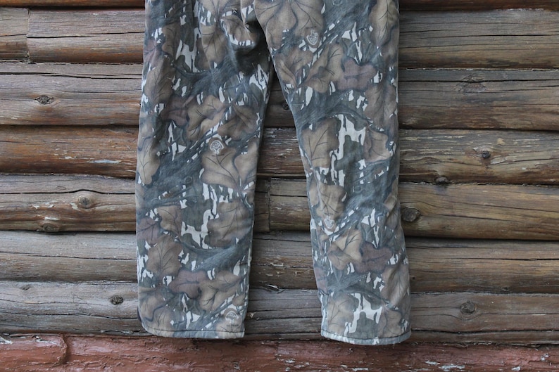 Puede incluir: Pantalones de camuflaje con un estampado de hojas en tonos marr&oacute;n, gris y blanco. Los pantalones est&aacute;n hechos de una tela duradera y son adecuados para actividades al aire libre. Los pantalones se muestran sobre un fondo de madera.
