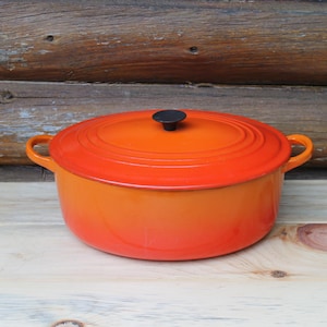 May include: An oval enameled cast iron Dutch oven featuring a vibrant gradient finish that transitions from a deep orange at the base to a lighter, bright orange on the lid. It includes two side handles and a small, round, black knob on the lid.