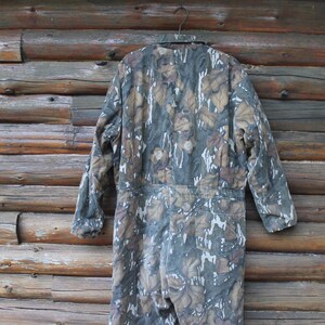 Puede incluir: Mono de camuflaje con un estampado de hojas en tonos verdes, marrones y blancos, colgado de una percha met&aacute;lica. La prenda de cuerpo entero se muestra sobre un fondo de troncos de madera. Tiene mangas largas y est&aacute; dise&ntilde;ado para actividades al aire libre.