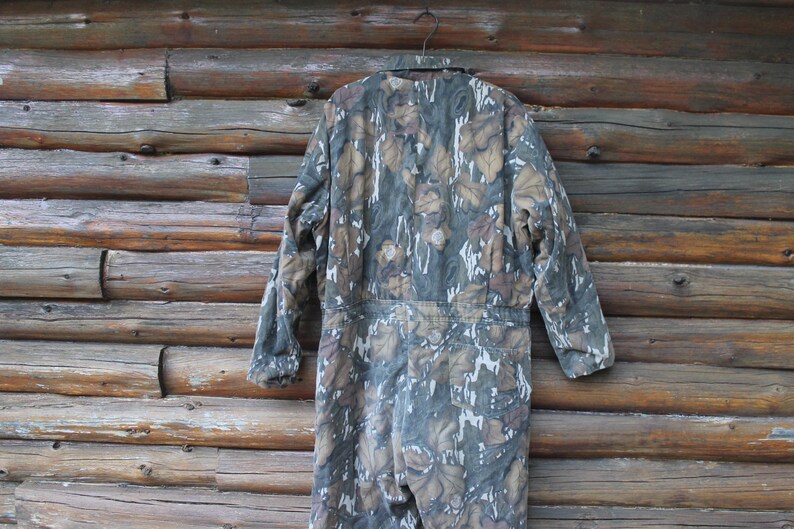 Puede incluir: Mono de camuflaje con un estampado de hojas en tonos verdes, marrones y blancos, colgado de una percha met&aacute;lica. La prenda de cuerpo entero se muestra sobre un fondo de troncos de madera. Tiene mangas largas y est&aacute; dise&ntilde;ado para actividades al aire libre.