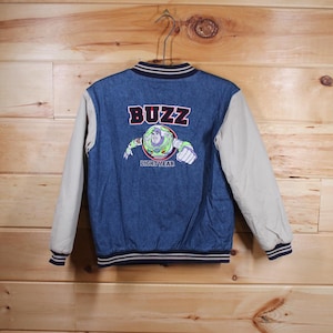 Puede incluir: Una chaqueta vaquera azul con una manga de estilo béisbol beige y blanca. La parte posterior de la chaqueta tiene una imagen bordada de Buzz Lightyear de la franquicia Toy Story con el texto "Buzz Lightyear".