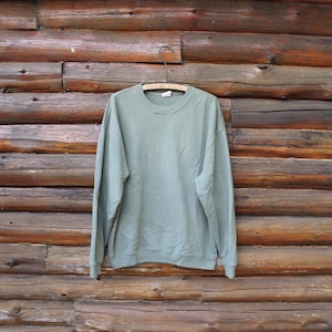 May include: A sage green sweatshirt with a crew neck and long sleeves hangs on a wooden hanger. The sweatshirt is made of a soft material and is displayed against a backdrop of weathered wooden logs.