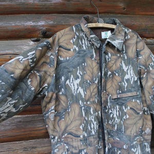 Puede incluir: Mono de camuflaje con un dise&ntilde;o de hojas marrones, grises y blancas. La prenda larga tiene cuello, cremallera frontal y bolsillo en el pecho. El mono est&aacute; colgado de una percha de madera sobre un fondo de madera.