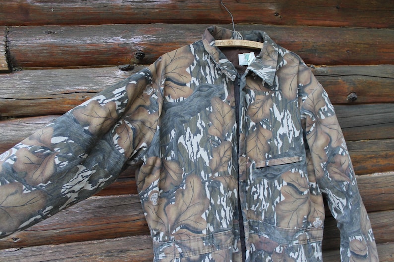 Puede incluir: Mono de camuflaje con un dise&ntilde;o de hojas marrones, grises y blancas. La prenda larga tiene cuello, cremallera frontal y bolsillo en el pecho. El mono est&aacute; colgado de una percha de madera sobre un fondo de madera.