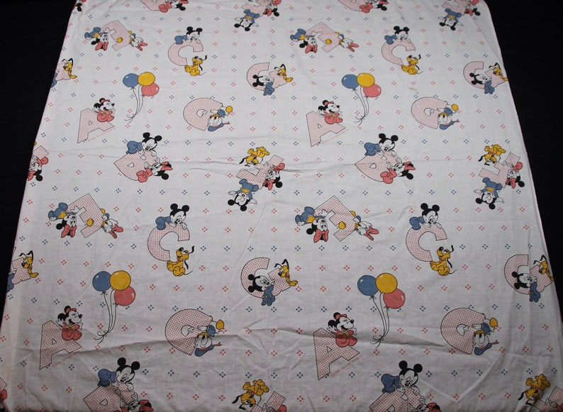 May include: A white baby blanket with a repeating pattern of Mickey Mouse, Minnie Mouse, Donald Duck, and Pluto. The characters are holding balloons and letters of the alphabet. The background is a light blue with small white dots.