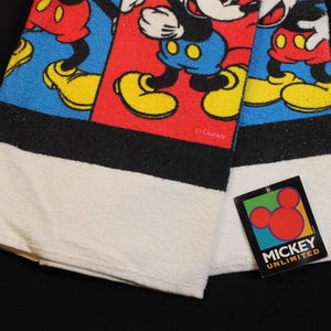 May include: A folded white and black towel featuring a colorful Mickey Mouse design. The design includes Mickey Mouse in red shorts, yellow shoes, and blue background. A tag with the words "Mickey Unlimited" is attached.