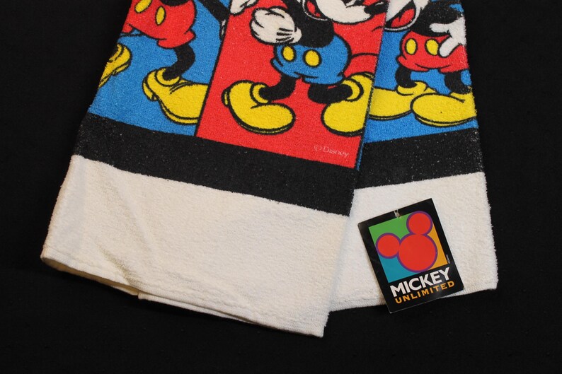 May include: A folded white and black towel featuring a colorful Mickey Mouse design. The design includes Mickey Mouse in red shorts, yellow shoes, and blue background. A tag with the words "Mickey Unlimited" is attached.