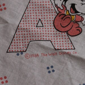 May include: A white fabric with a red and white polka dot letter A and a red cartoon character. The text "&copy;1984 The Walt Disney Co." is printed below the letter.