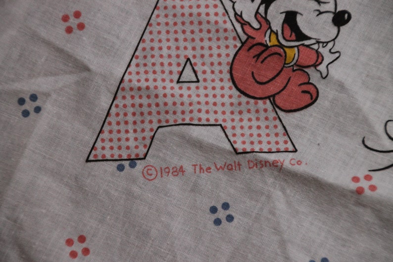 May include: A white fabric with a red and white polka dot letter A and a red cartoon character. The text "&copy;1984 The Walt Disney Co." is printed below the letter.