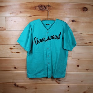 Pode incluir: Uma camisa de beisebol turquesa com mangas curtas e gola em V. A camisa apresenta a palavra "Riverwood" em escrita preta no peito. A camisa está abotoada e pendurada em um cabide de madeira contra um fundo de painéis de madeira.