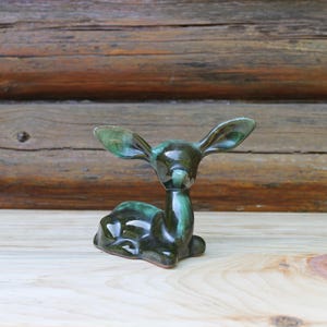 May include: A ceramic deer figurine in a reclining pose, glazed in shades of green. The deer has large, upright ears and a smooth, glossy finish. The figurine is set against a wooden background.
