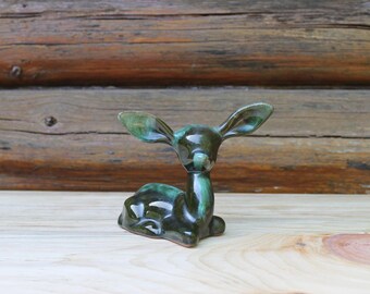 May include: A ceramic deer figurine in a reclining pose, glazed in shades of green. The deer has large, upright ears and a smooth, glossy finish. The figurine is set against a wooden background.
