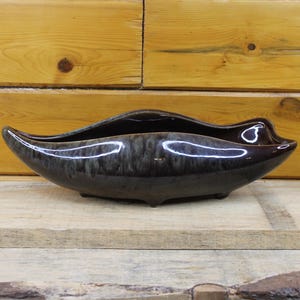 May include: A dark brown ceramic planter with a glossy finish. The elongated, boat-shaped vessel has a wavy rim and sits on three small feet. The interior is a darker shade, creating a contrast with the exterior.
