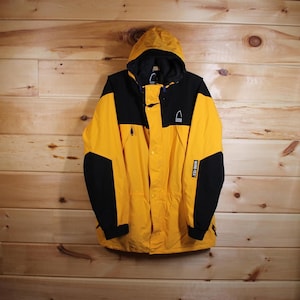 Puede incluir: Chaqueta con capucha amarilla y negra con el texto "GORE-TEX" en la manga. La chaqueta tiene una capucha negra y paneles negros en los hombros y las mangas. La chaqueta tiene un logotipo en el pecho.