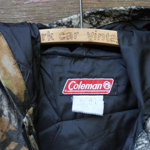 May include: A camouflage-patterned jacket with a black interior lining. The jacket features a red and white "Coleman" brand label. The jacket is hanging on a wooden hanger with the text "rk car vintage".