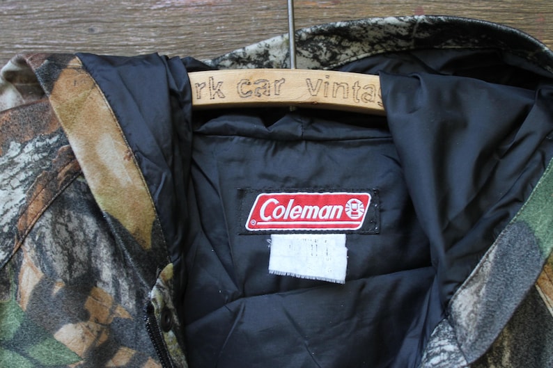 May include: A camouflage-patterned jacket with a black interior lining. The jacket features a red and white "Coleman" brand label. The jacket is hanging on a wooden hanger with the text "rk car vintage".