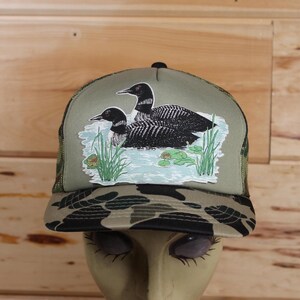 May include: A green camouflage trucker hat with a black and white embroidered image of two loons swimming in a lake with lily pads and reeds.